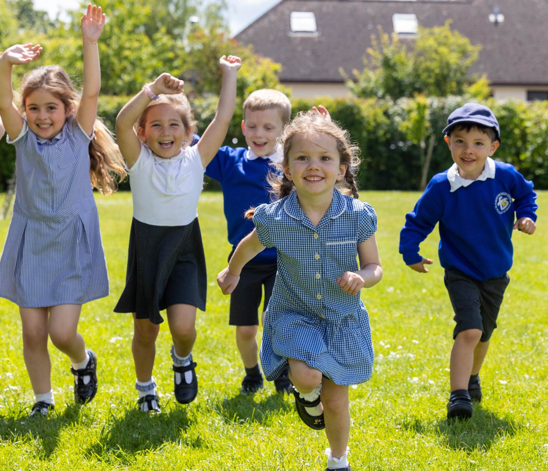 a group of young primary school wearing blue check, white and blue uniform are running across a field in the summer. They are smiling and raising their arms in celebration