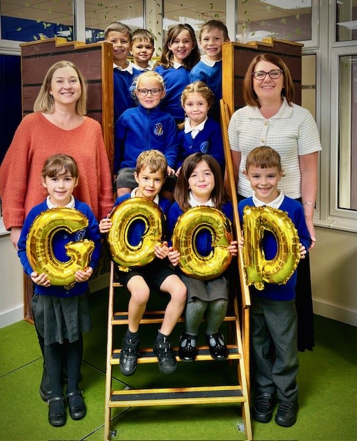 image of  schoolchildren wearing blue unform with two women . The children are holding up gold foil balloons that say GOOD