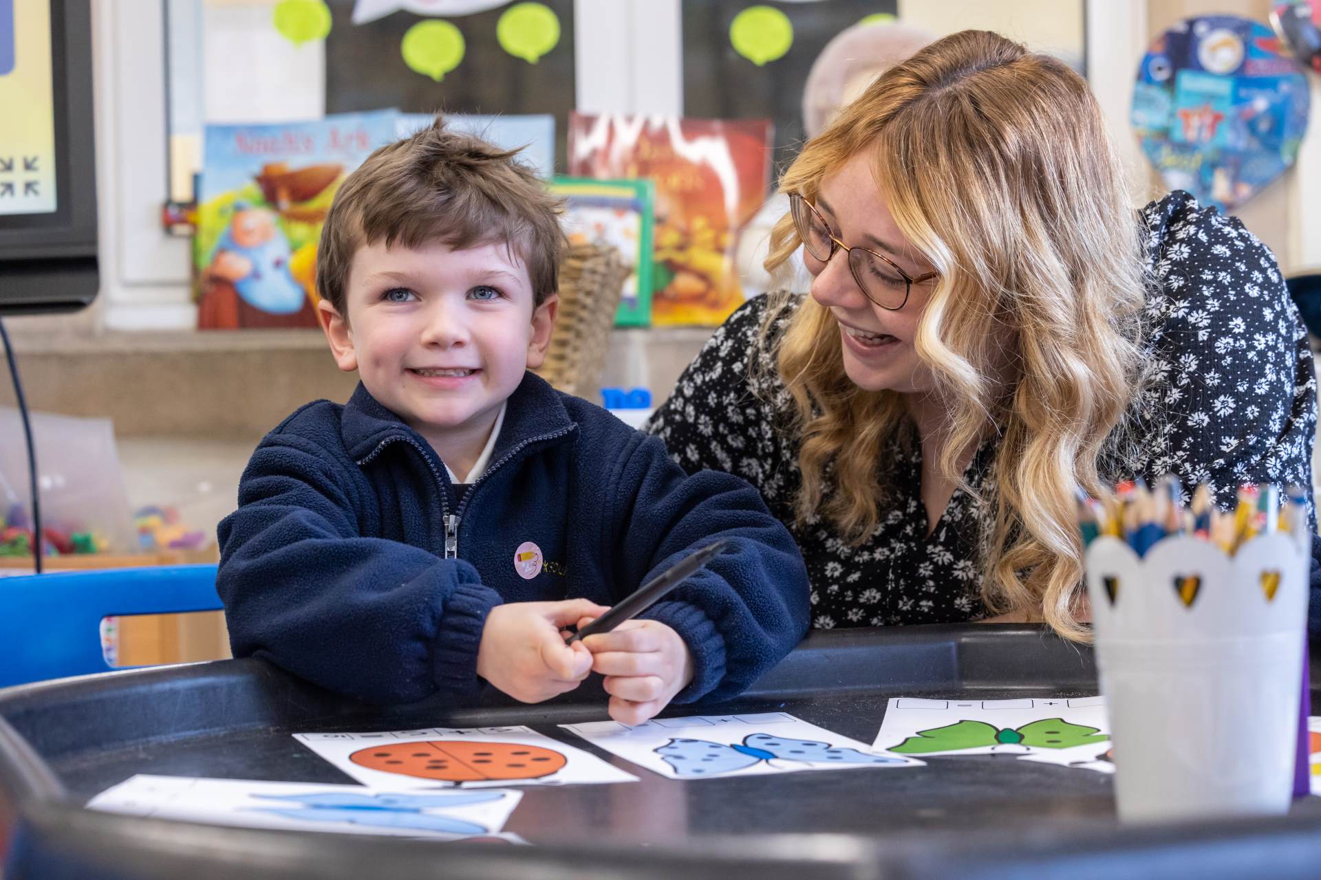 a young boy in a blue jumper sat in a classroom with his female teacher. They are both smiling