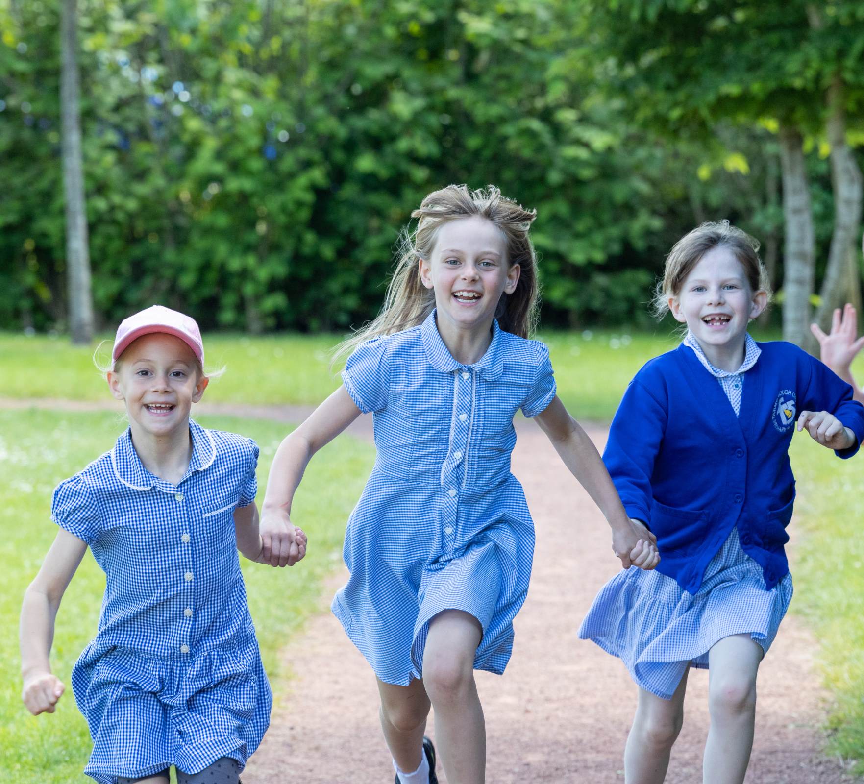 three girls wearing blue school uniform are running towards the camera and smiling. In the background is a green field and green trees. The caption reads Pupils are proud to belong to their school