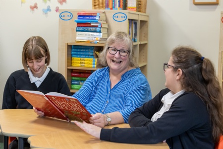 two young female students from Fosse Way Special schoo are looking at a book with an older woman and smiling
